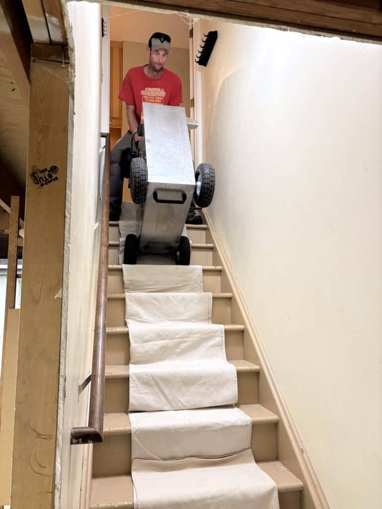 Our Drain and Sewer technician carrying a flex-shaft milling machine down tarp-covered stairs