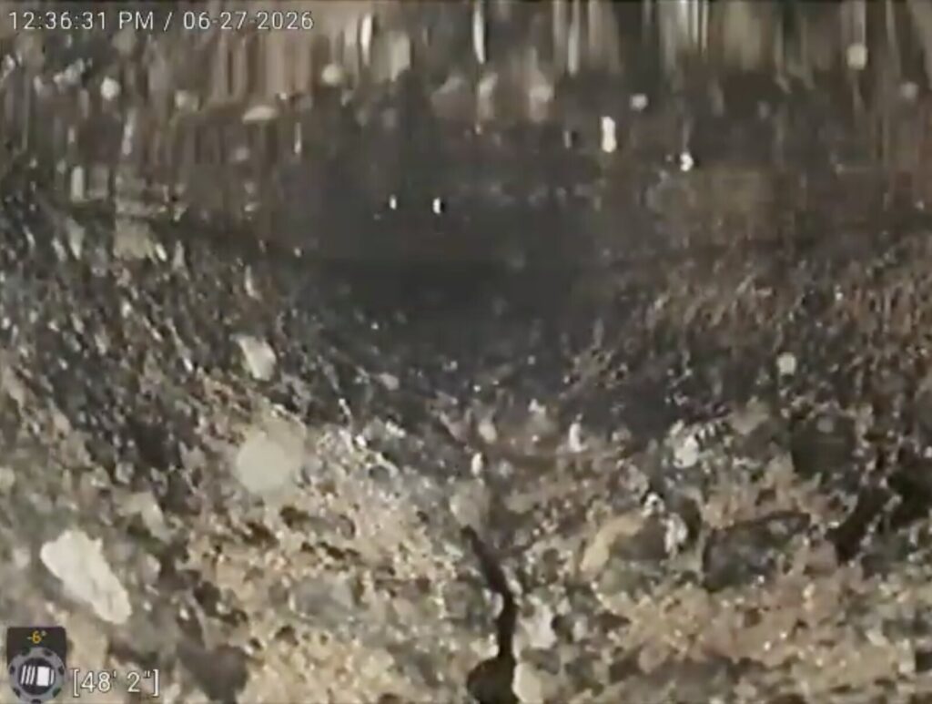 Close view of a concrete sewer pipe wall with individual stones standing proud of the surface above a dark eroded channel.